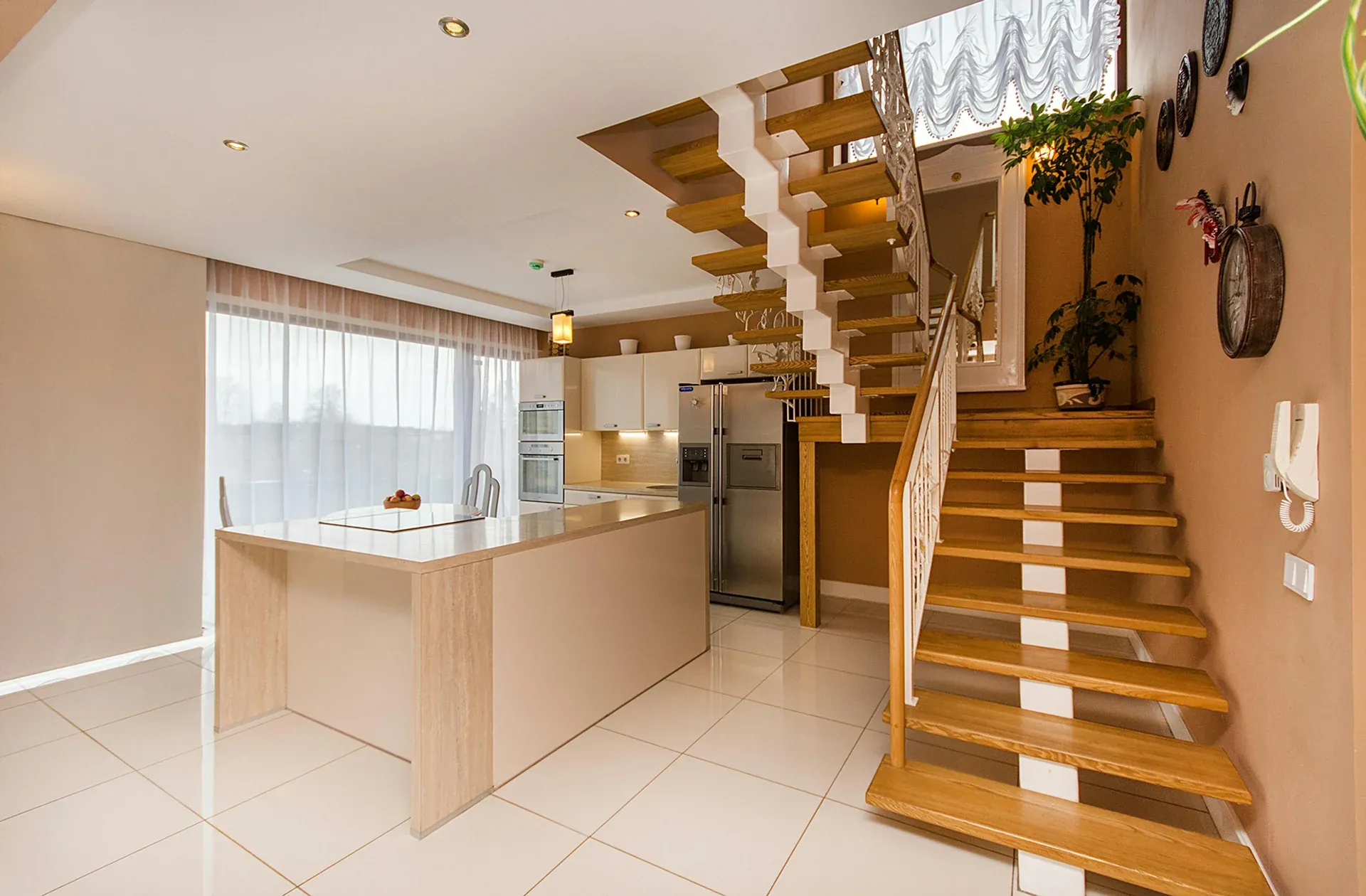 A modern open-plan kitchen and living space featuring a white kitchen island, integrated appliances, and a light wooden floating staircase.