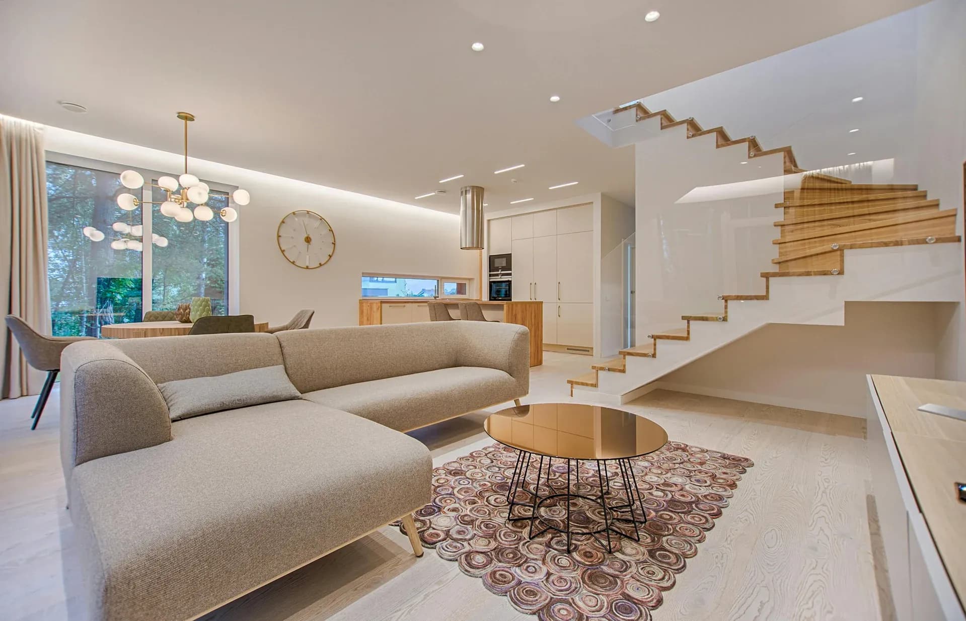 Modern open-plan living room and kitchen featuring a large sectional sofa, coffee table, dining area, a contemporary kitchen with island, and a stylish wooden staircase with glass balustrade.