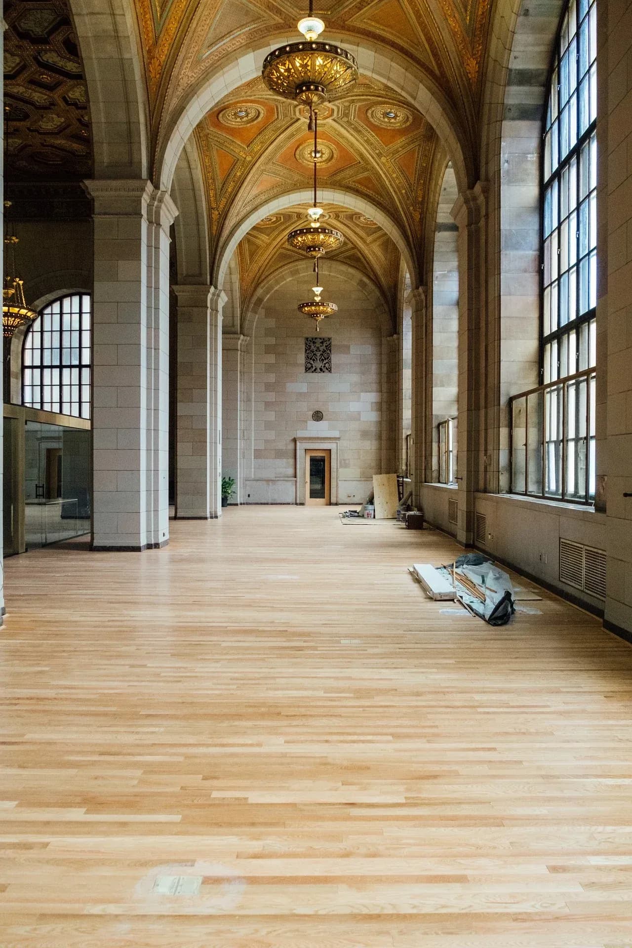 A grand, empty hall with newly installed light wooden flooring, tall stone pillars, large arched windows, and ornate gold and orange vaulted ceilings.
